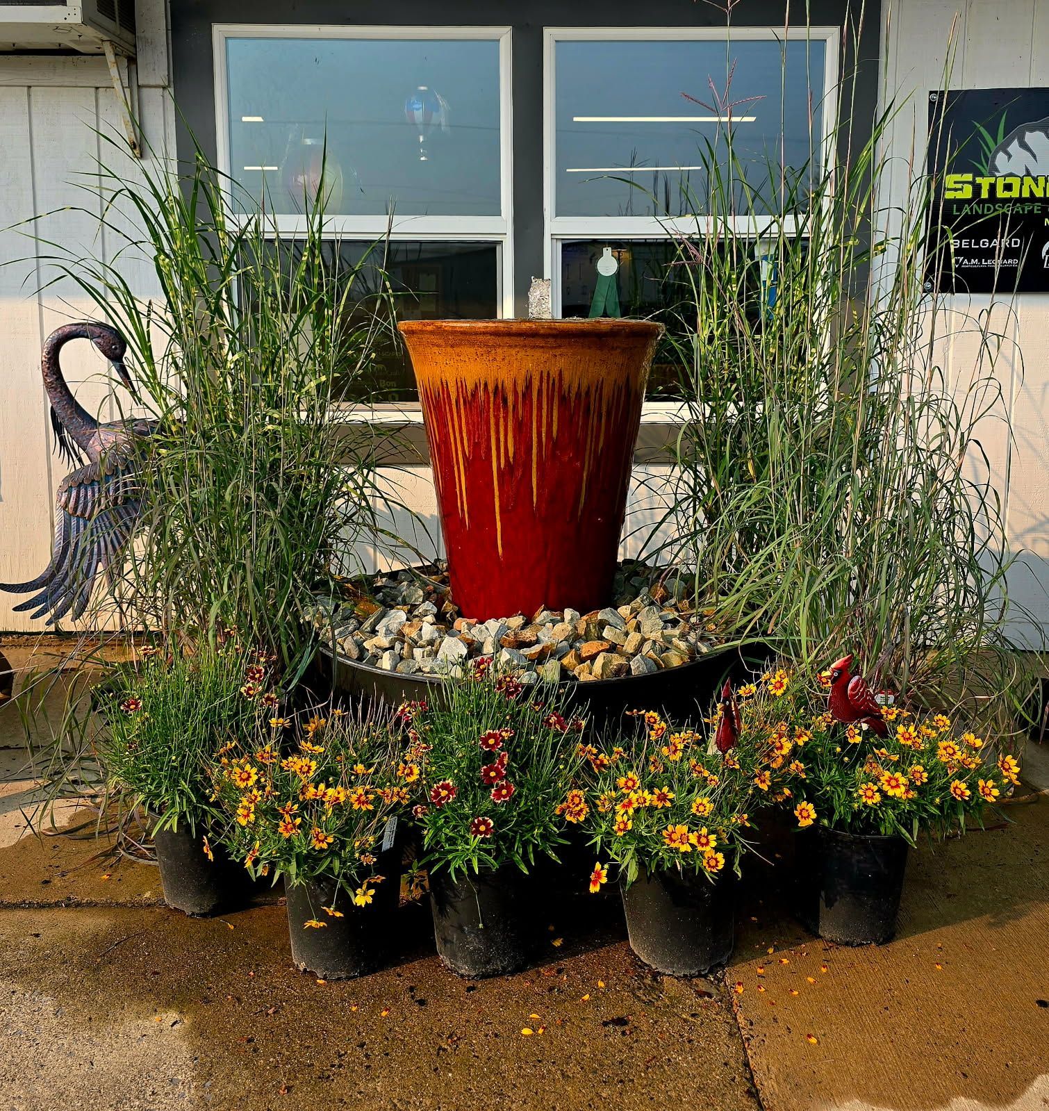 Decorative potted flowers and grasses around a tall red planter in front of a storefront window