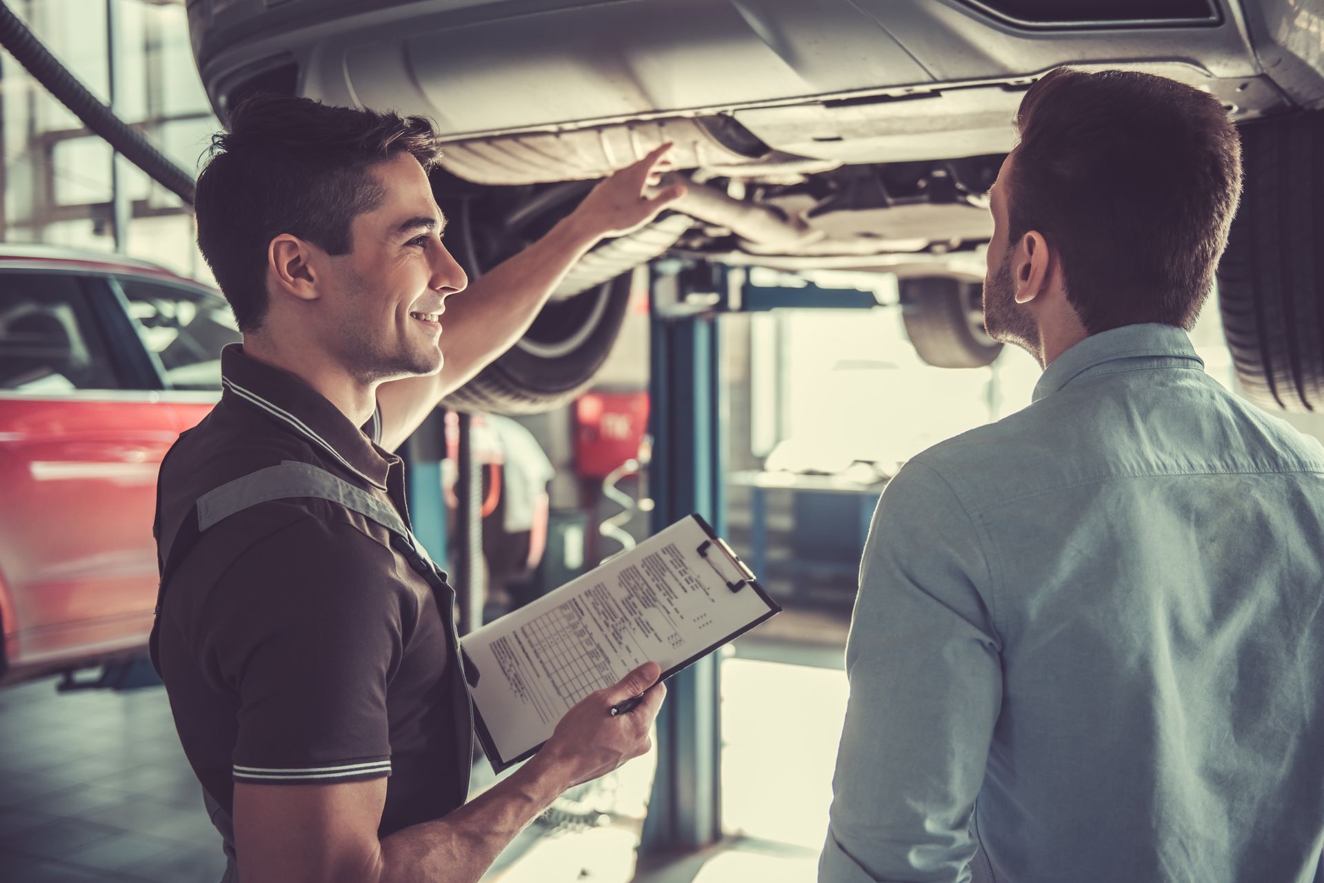 Two mechanics talking under a raised car in an auto repair shop, one holding a clipboard
