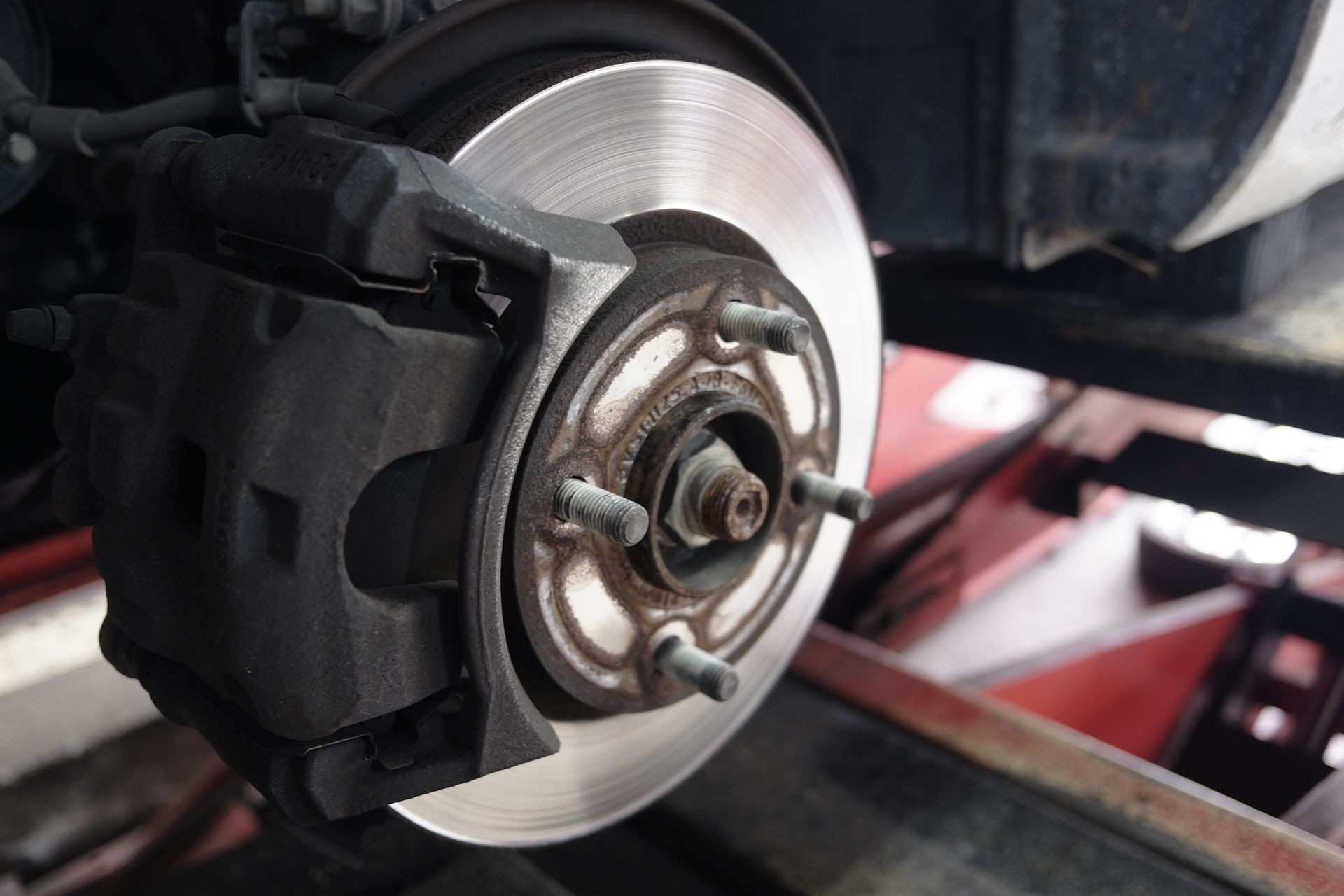 Close-up of a car brake rotor and caliper mounted on a lift inside an auto repair shop.