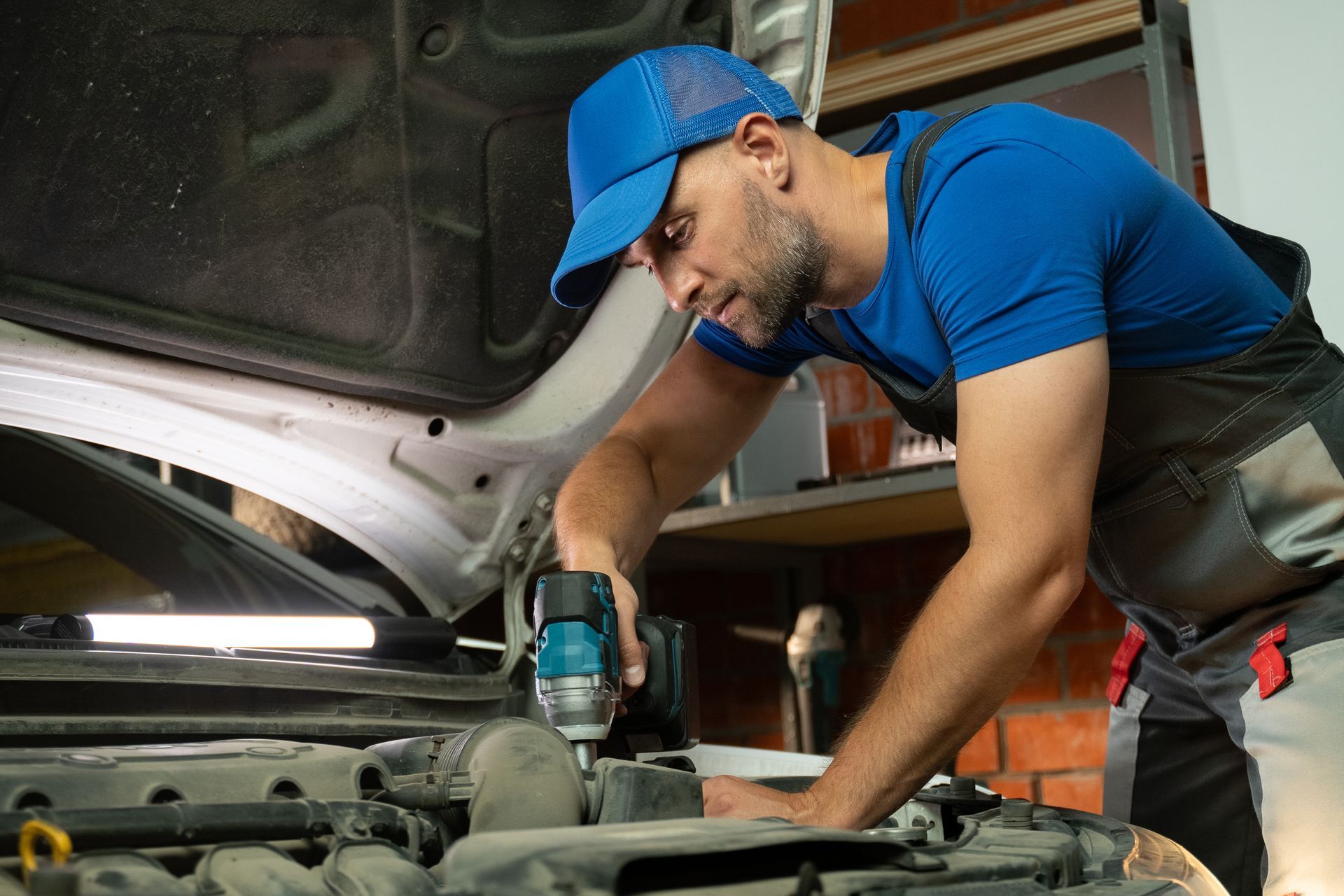 A mechanic works on a car engine in a garage. He is using a power tool to tighten bolts and screws. Mechanic in blue cap using a power tool under a car hood in a garage