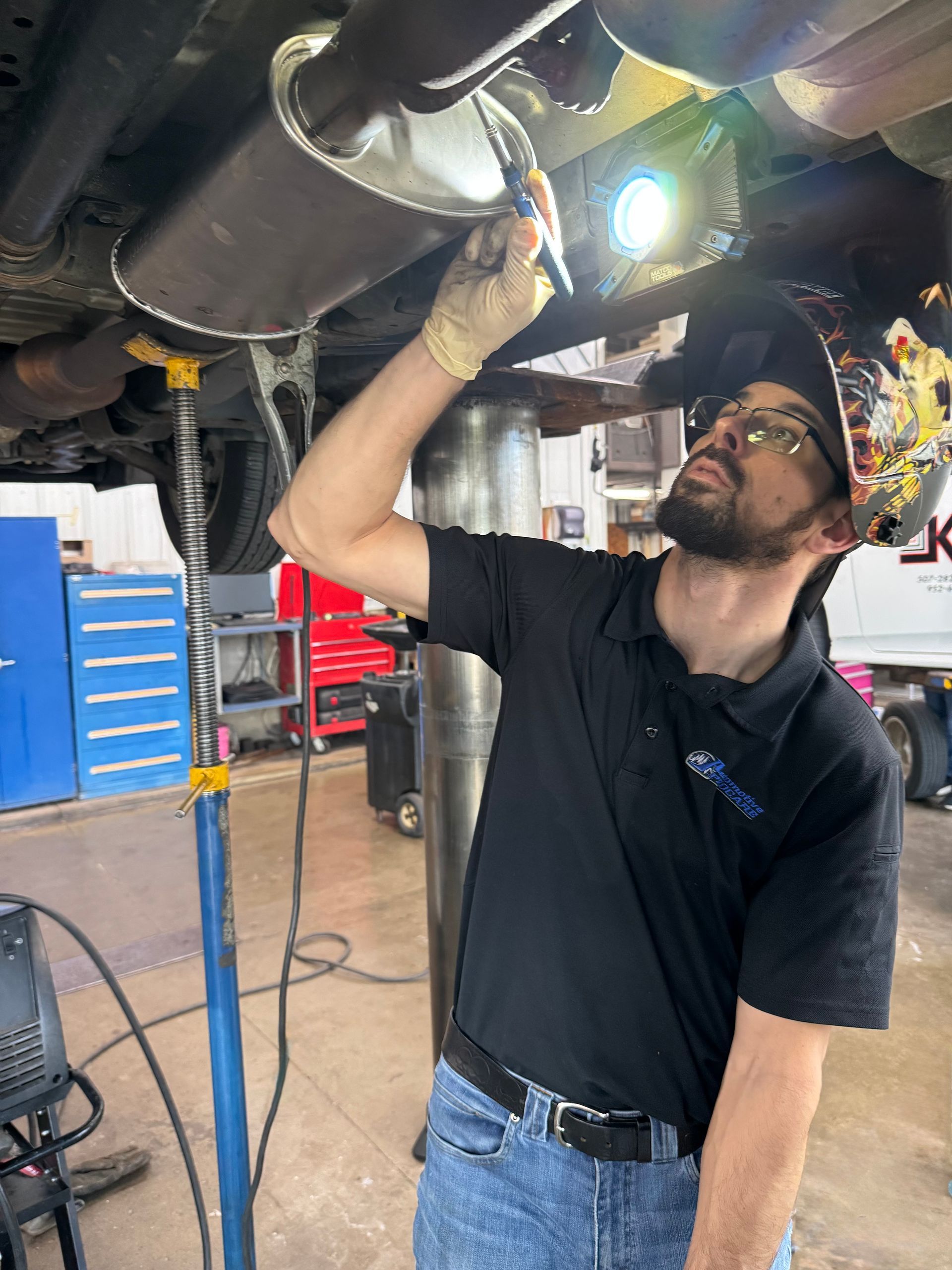 Mechanic inspecting a vehicle underside in a garage, using a flashlight and wearing gloves.