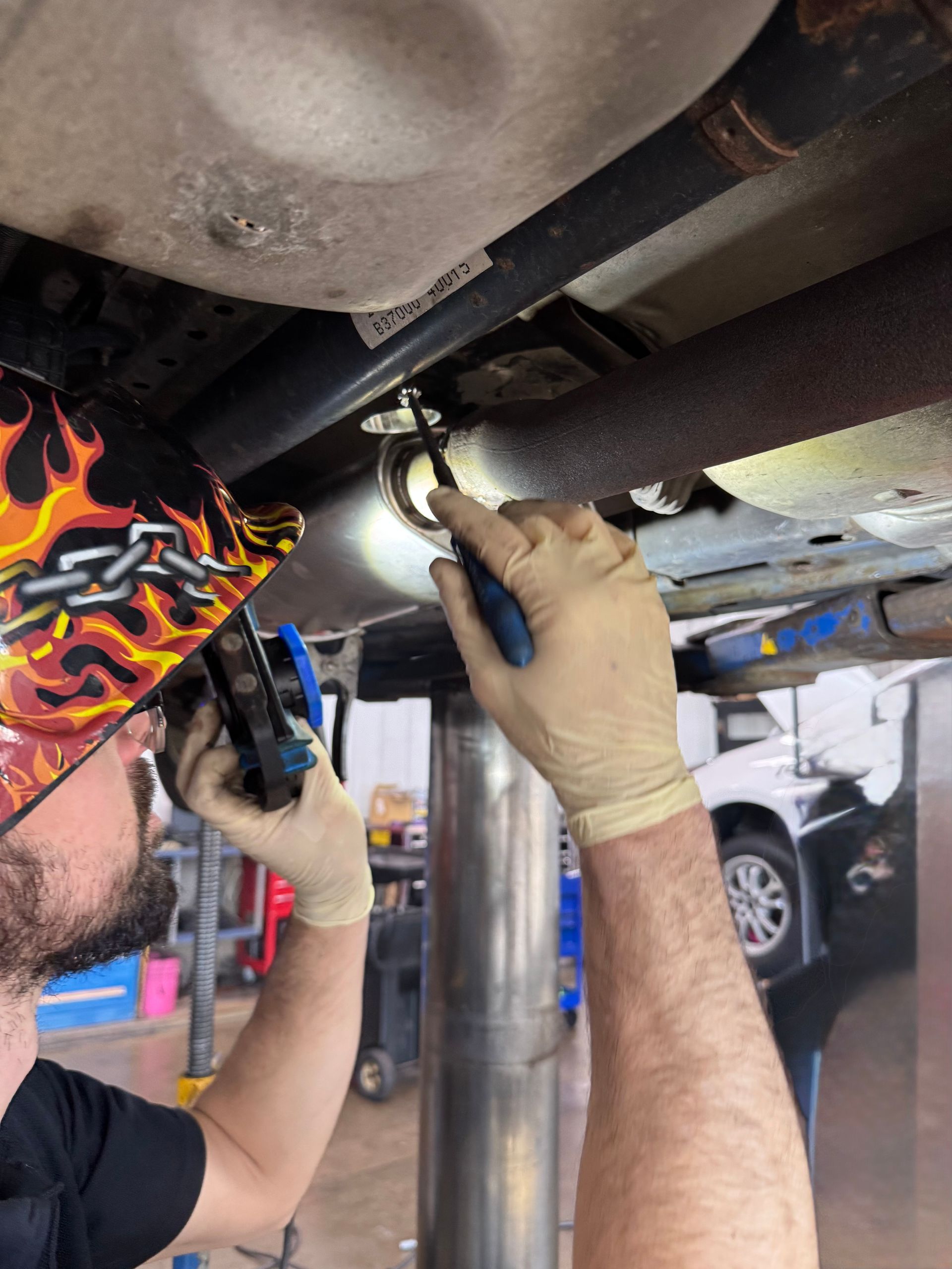 Mechanic tightening a bolt under a vehicle with a wrench in a garage, wearing gloves and a cap.