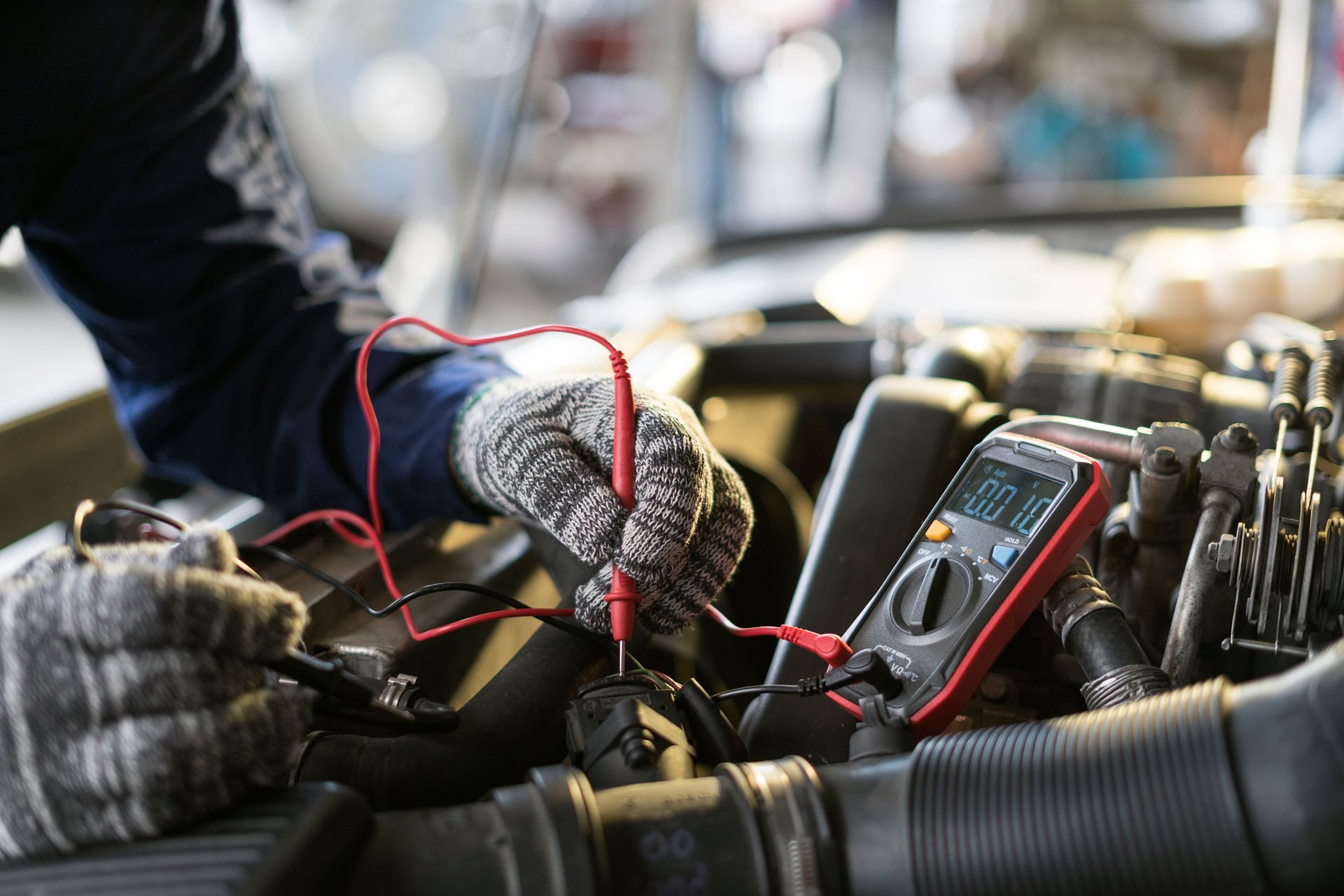 Gloved hands testing car engine wiring with a multimeter in a workshop.