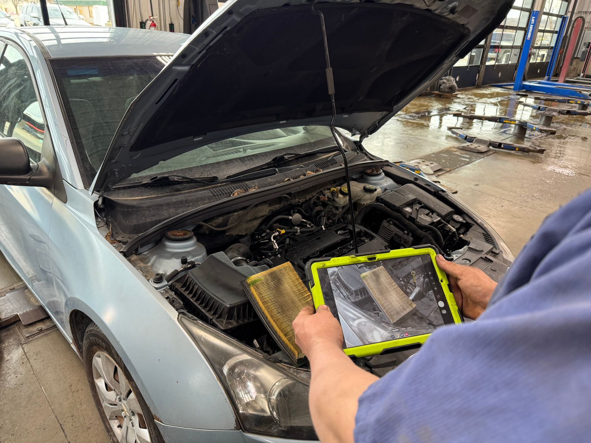 Person holding a yellow-framed air filter over an open car hood in a repair shop