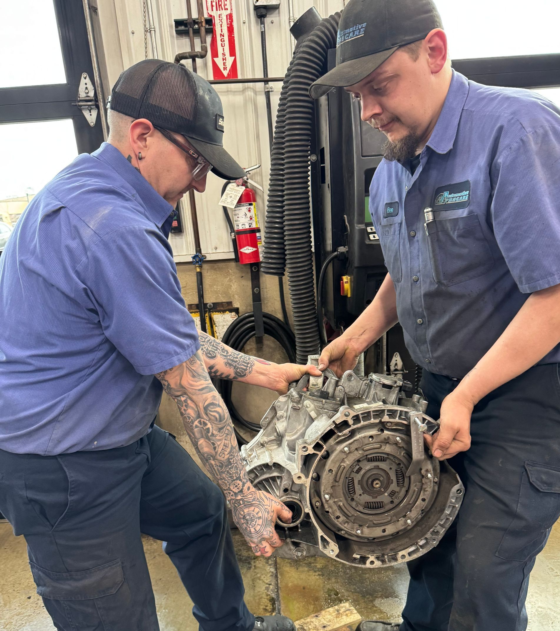 Two mechanics in blue uniforms handling a large transmission in an auto shop