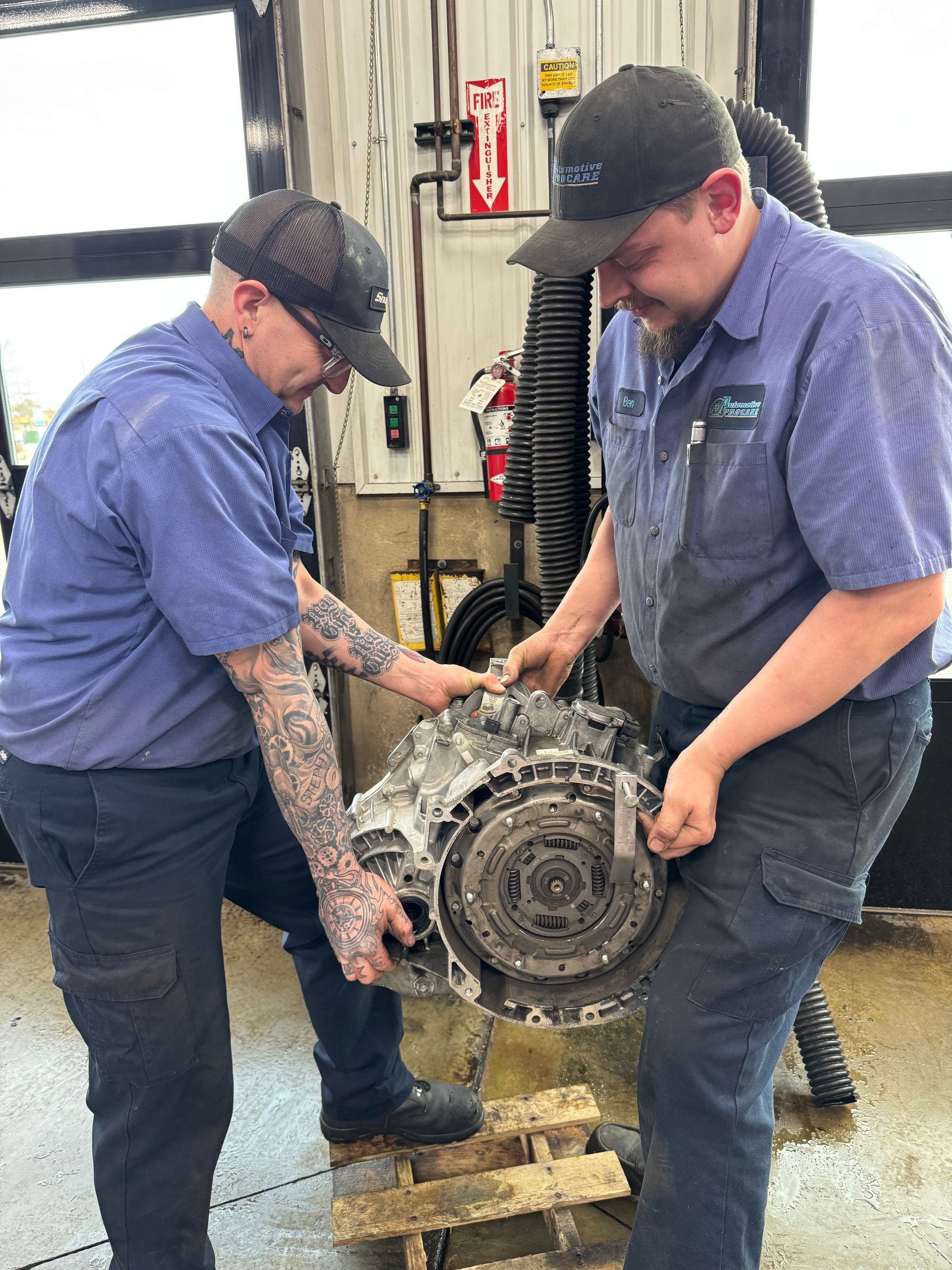 Two mechanics in blue shirts lift a large engine or transmission in an auto shop.