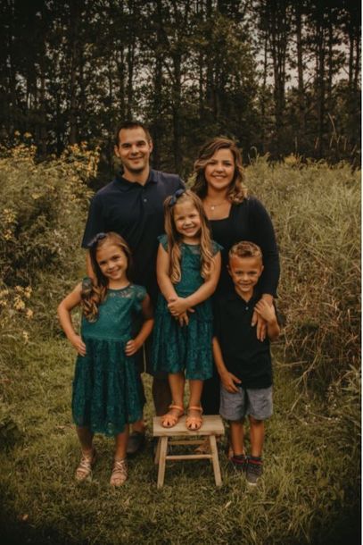 Family posing outdoors in a grassy field, with four children in teal outfits and two adults behind them