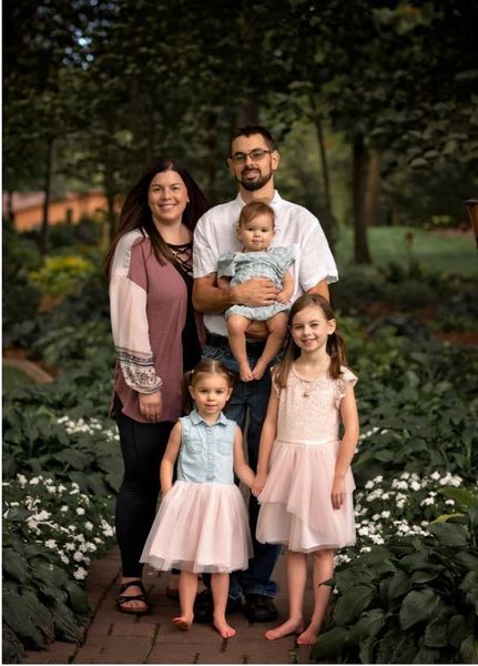 Family of four standing in a garden, smiling with two children in pastel dresses.