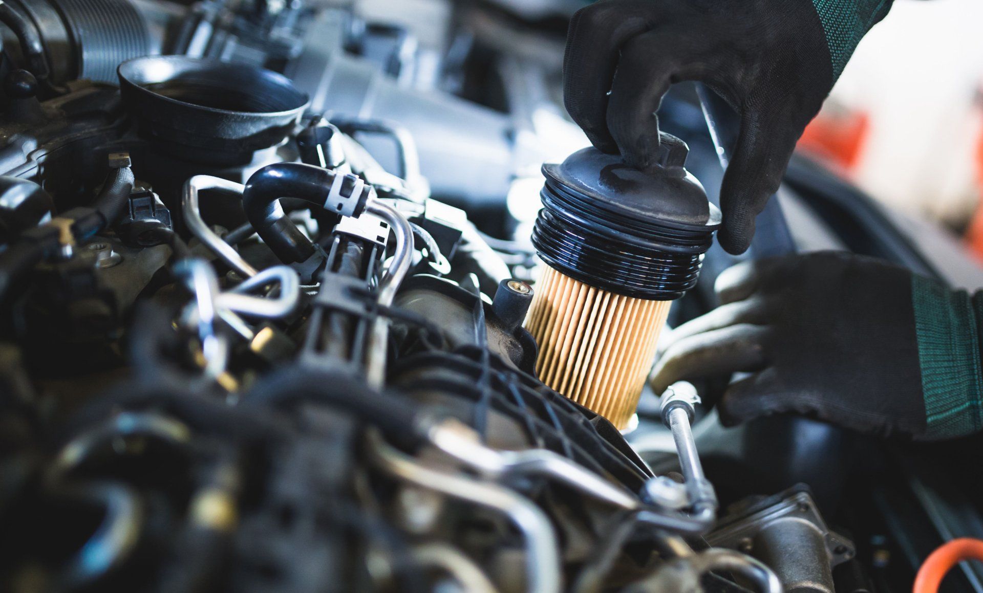 Gloved hands inspect a car engine with a yellow oil filter in a garage