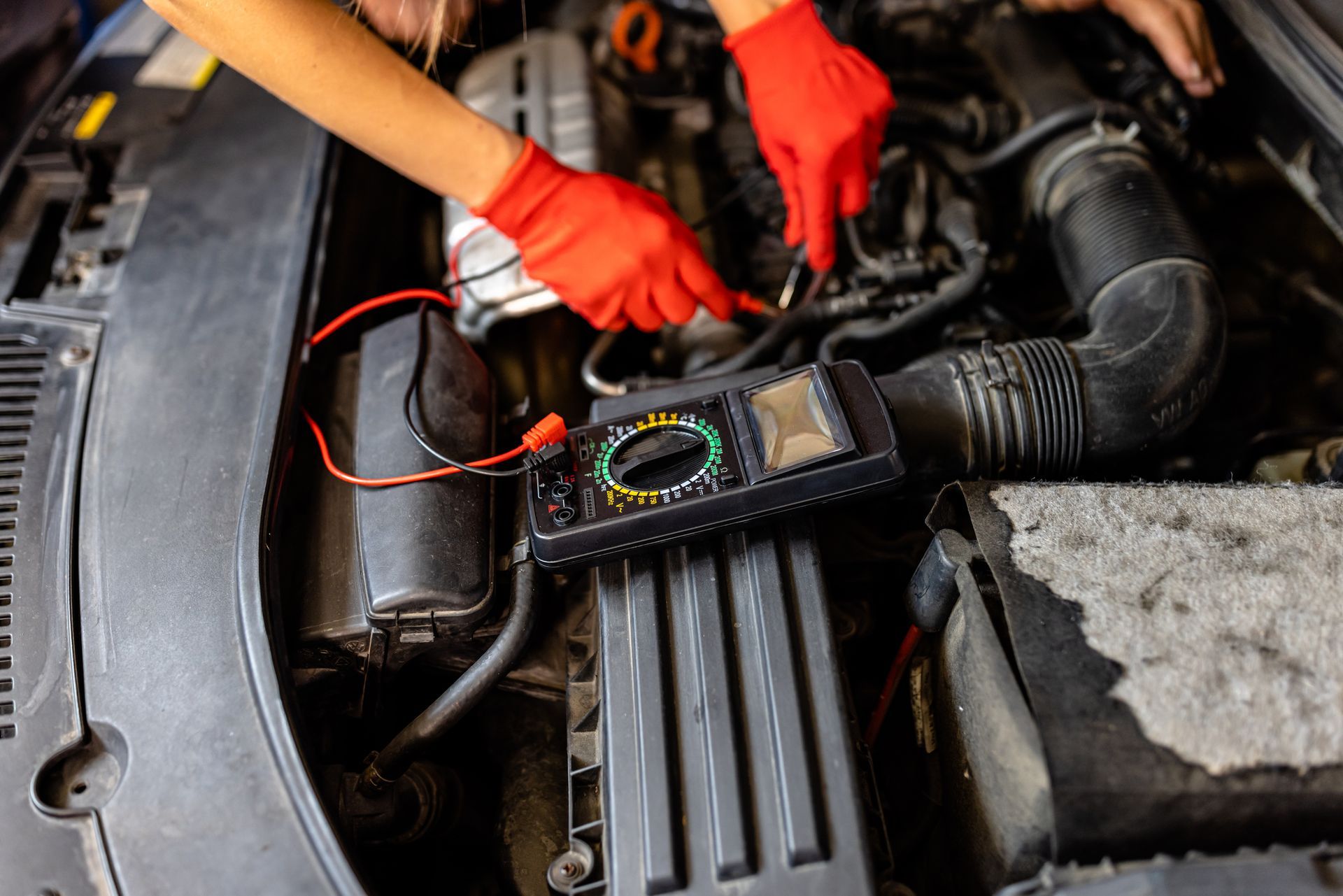 Mechanic using a multimeter with red-gloved hands to test wiring in a car engine bay