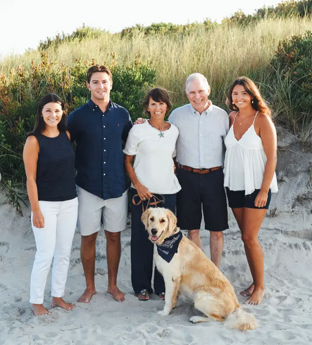 Group of five adults and a dog posing on a sandy beach before dune grass.