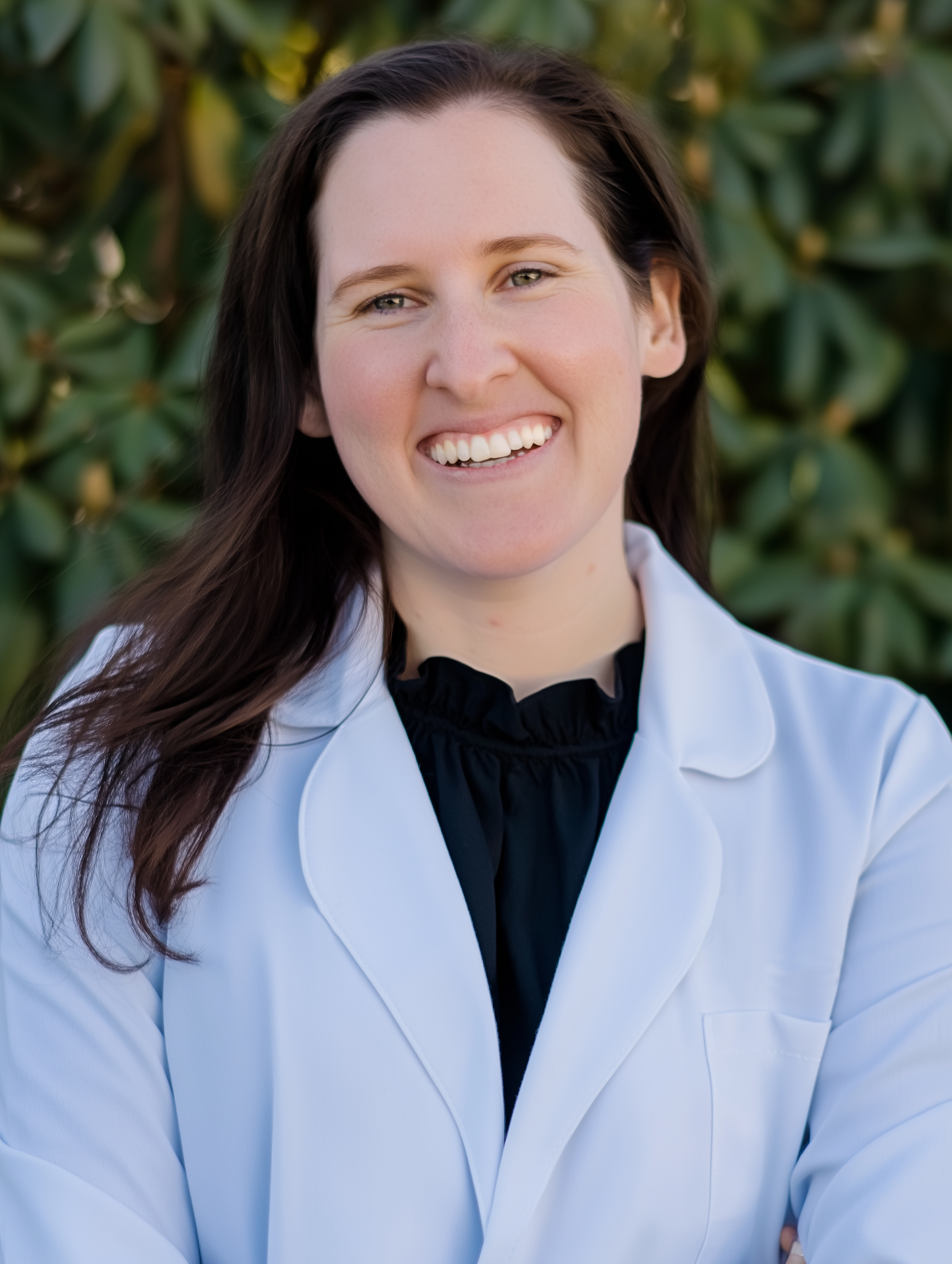 Woman in white coat smiles outdoors; dark hair, green background.