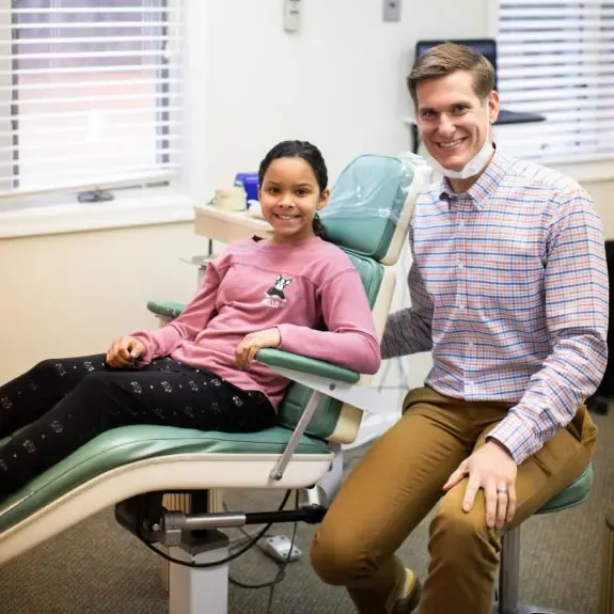 Young girl smiles in a dentist chair next to a smiling male dentist in a light-filled office.