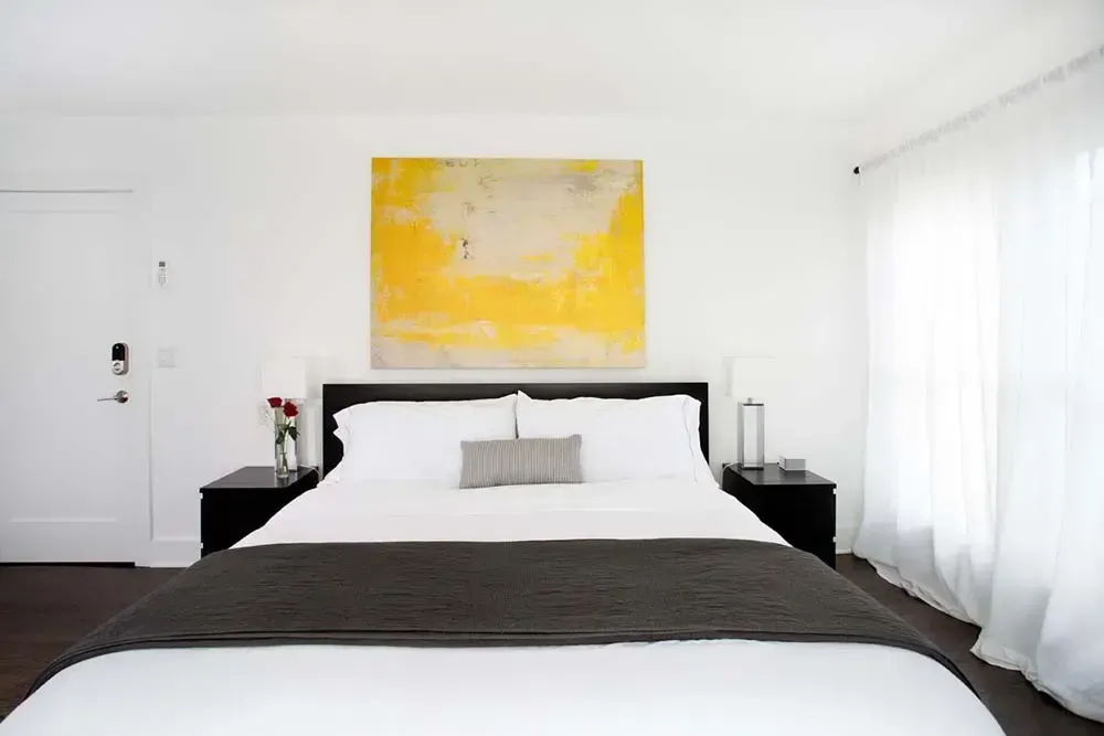 Bedroom with white walls, king bed, yellow artwork, and dark nightstands.