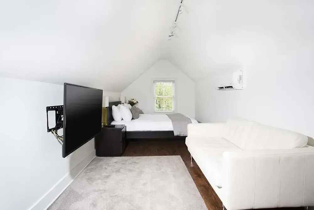 White bedroom with TV, bed, and white sofa. Wooden floor, angled white ceiling.