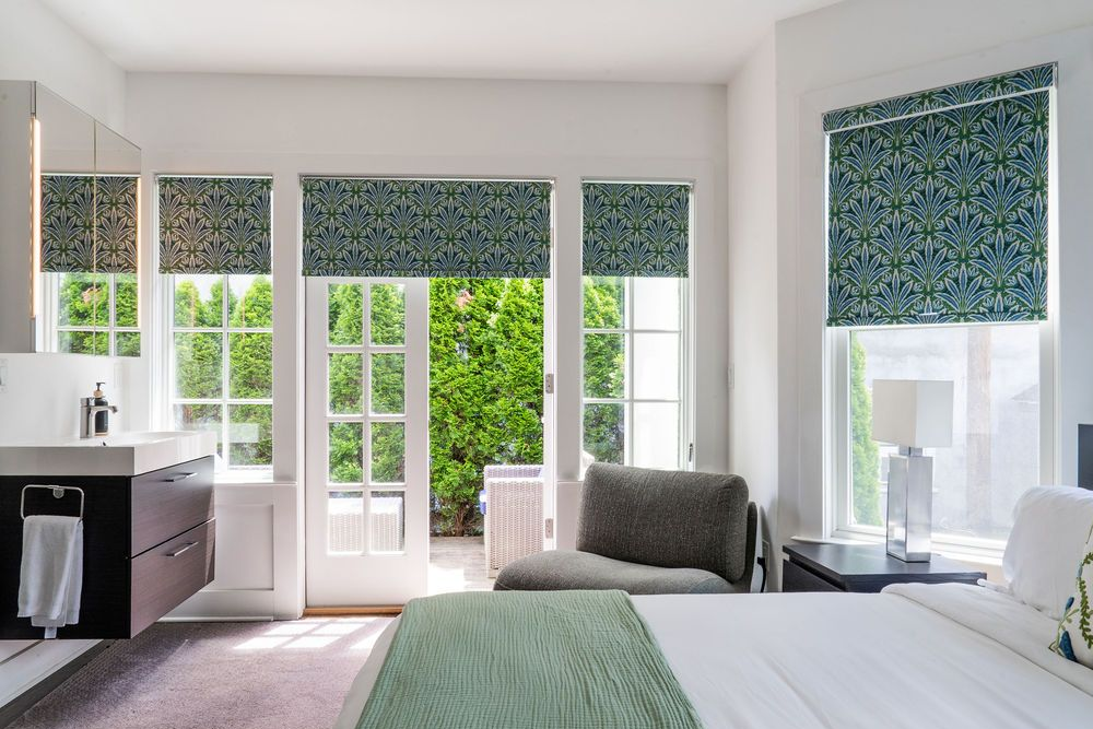Bedroom with white curtains, gray bed, and a small table with a lamp and flowers.