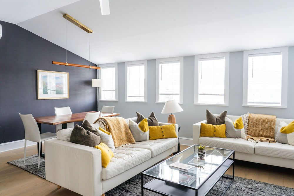 Modern living room with white sofas, dining table, dark accent wall, and multiple windows.