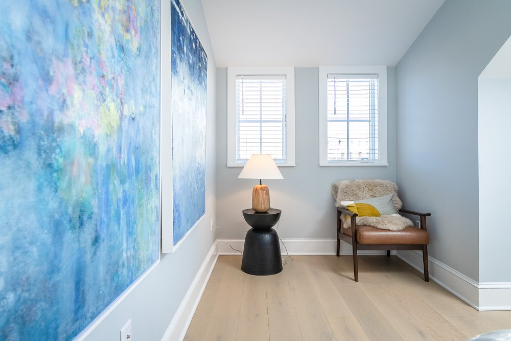 Cozy reading nook with art, windows, a chair, and a lamp on a side table. Light blue walls, wood floor.