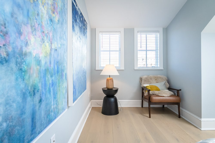 Cozy reading nook with art, windows, a chair, and a lamp on a side table. Light blue walls, wood floor.