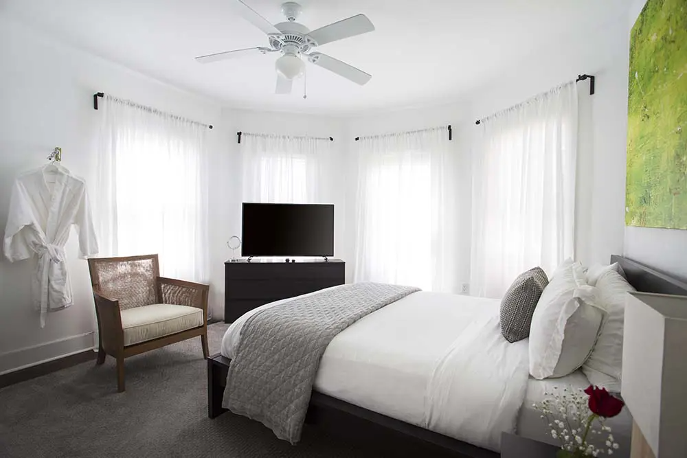 Bedroom with bed, TV, chair, windows with white curtains, and a ceiling fan.