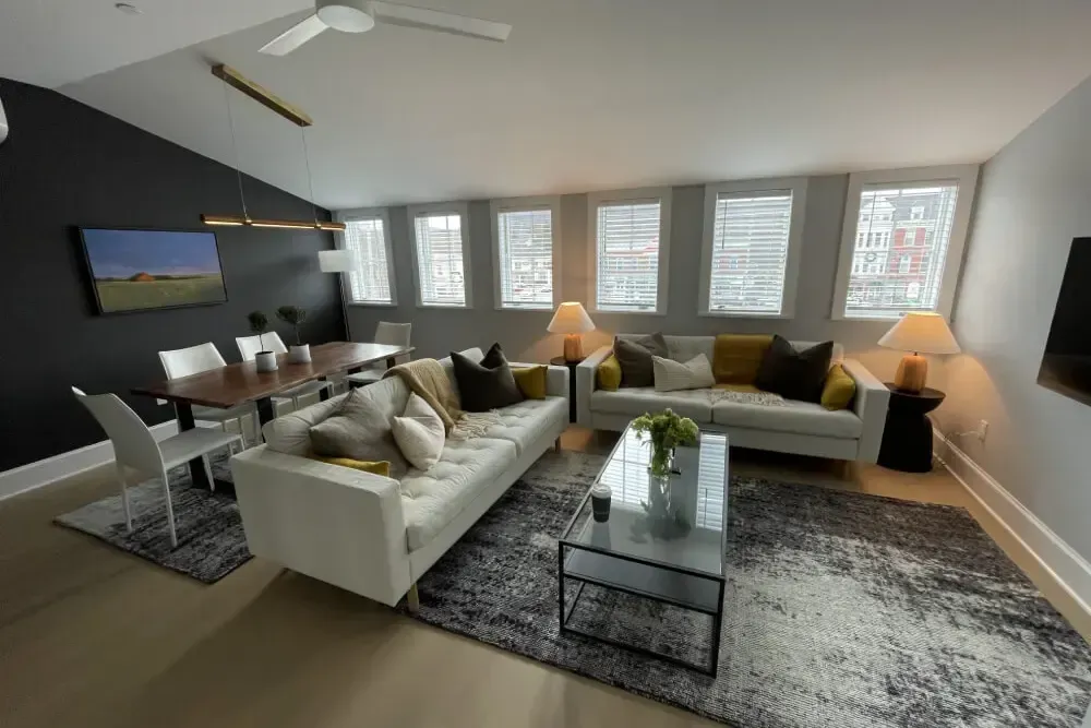 Modern living room with white sofas, dining table, dark accent wall, and multiple windows.