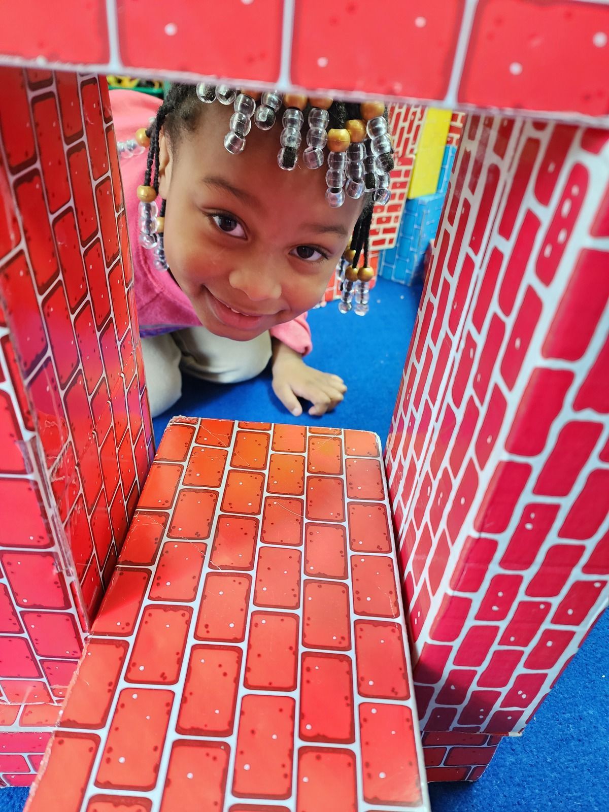 Child peeking through a red brick-pattern play structure, smiling at the camera