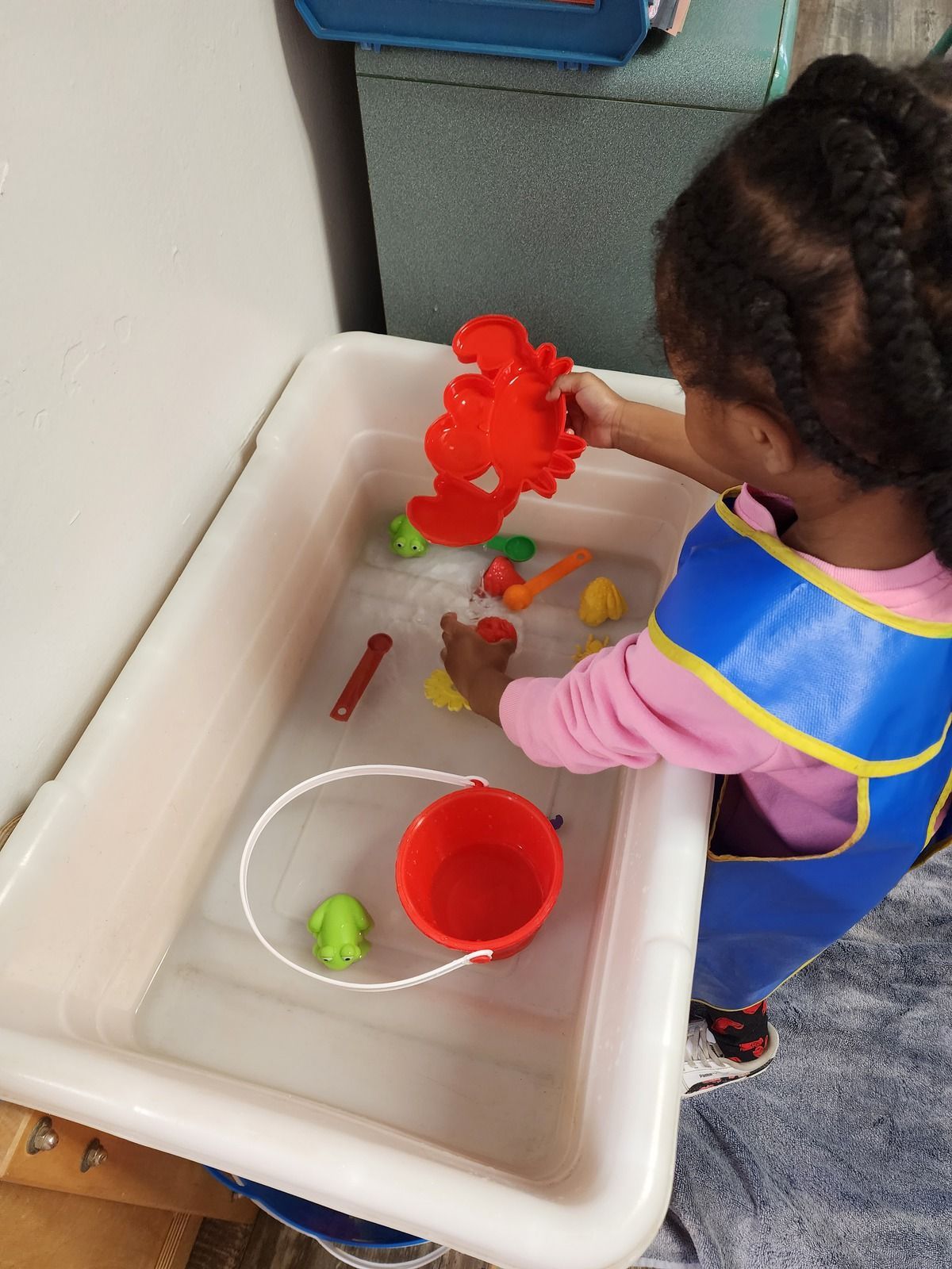Child playing with water toys in a white bin, using a red scoop and colorful floating toys.
