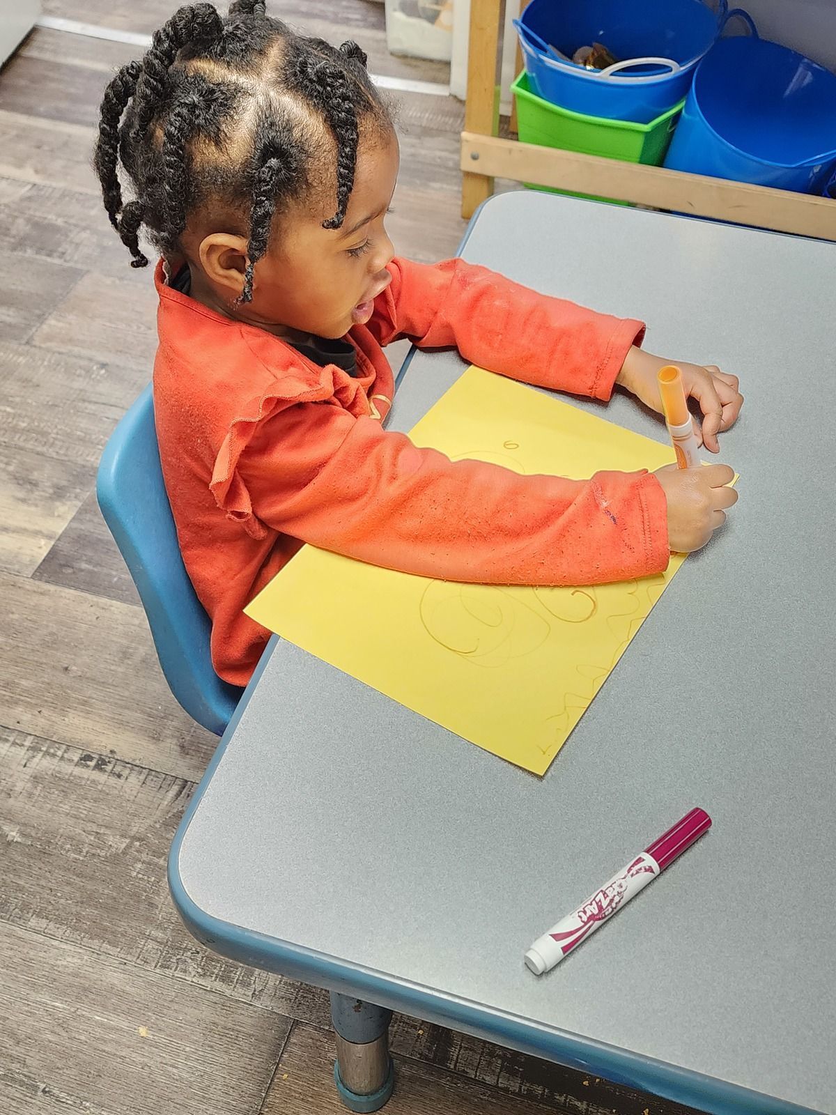 Child in orange shirt drawing on yellow paper at a classroom table