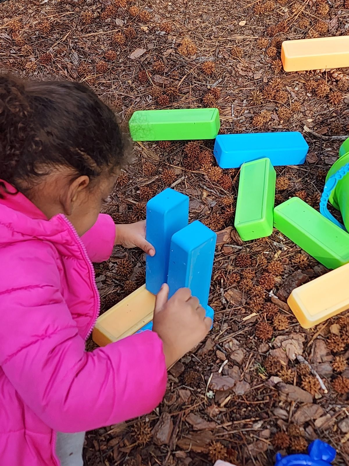 Child in a pink jacket building with colorful blocks on the ground outdoors.