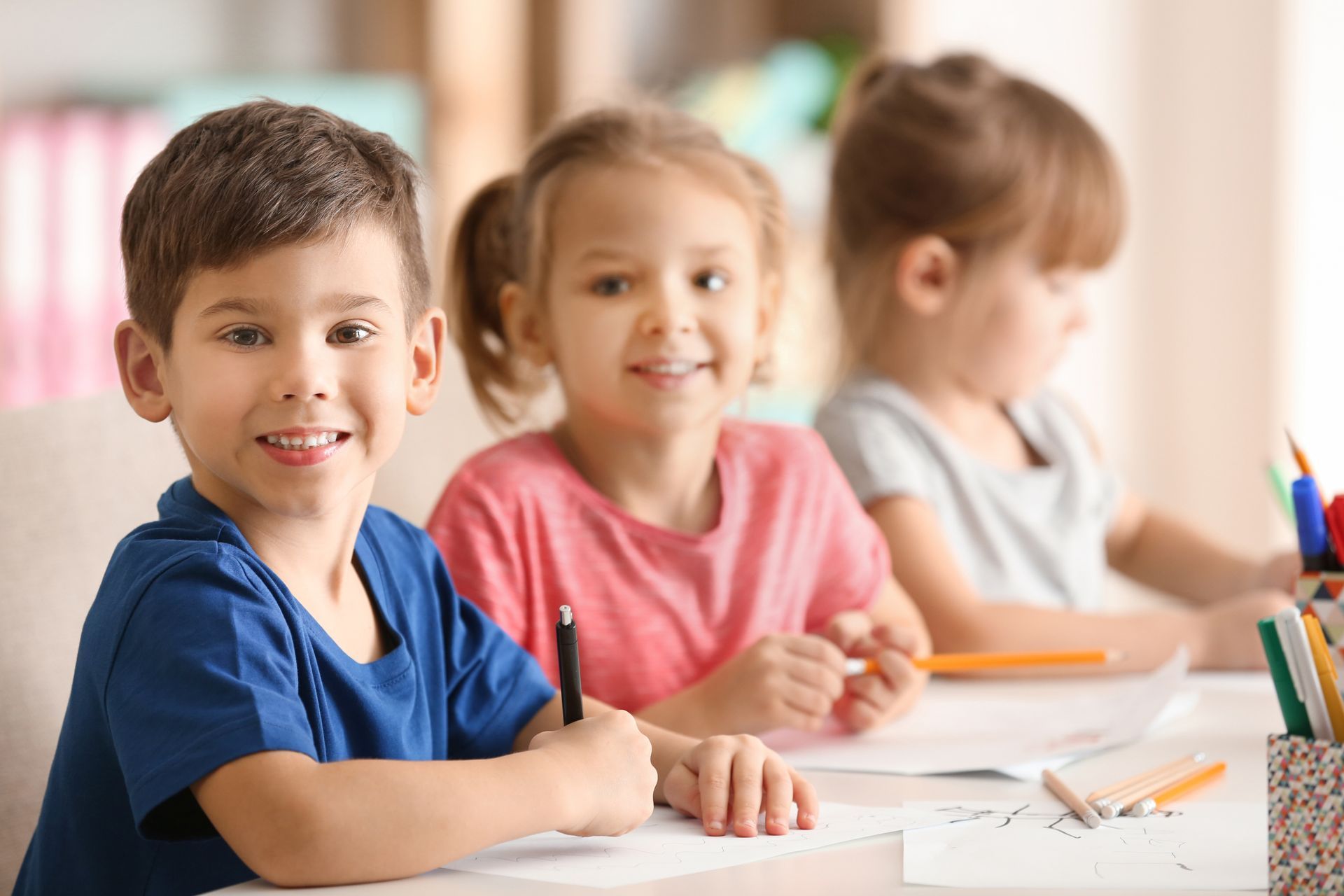 Three children sit at a classroom table, smiling and writing with pencils.