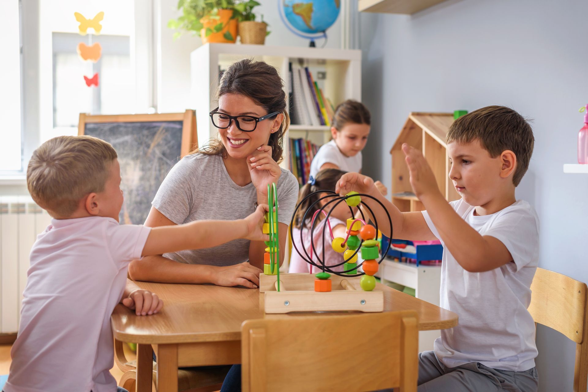 Children and a teacher play with a colorful bead maze at a classroom table.