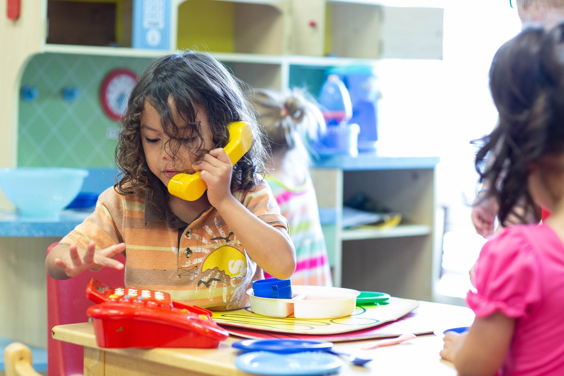 Child playing with a yellow toy phone at a colorful table, with another child nearby and toys around.