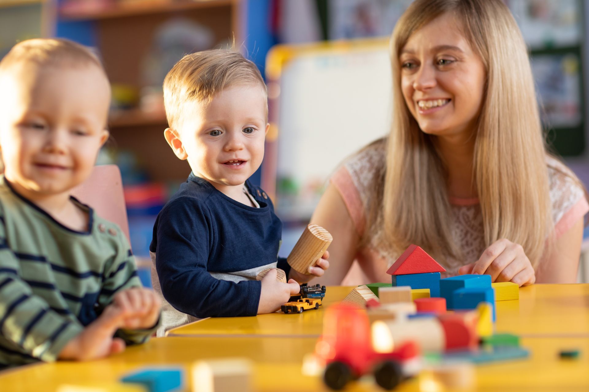 Two children and an adult smiling at a colorful table with toys and blocks in a playroom
