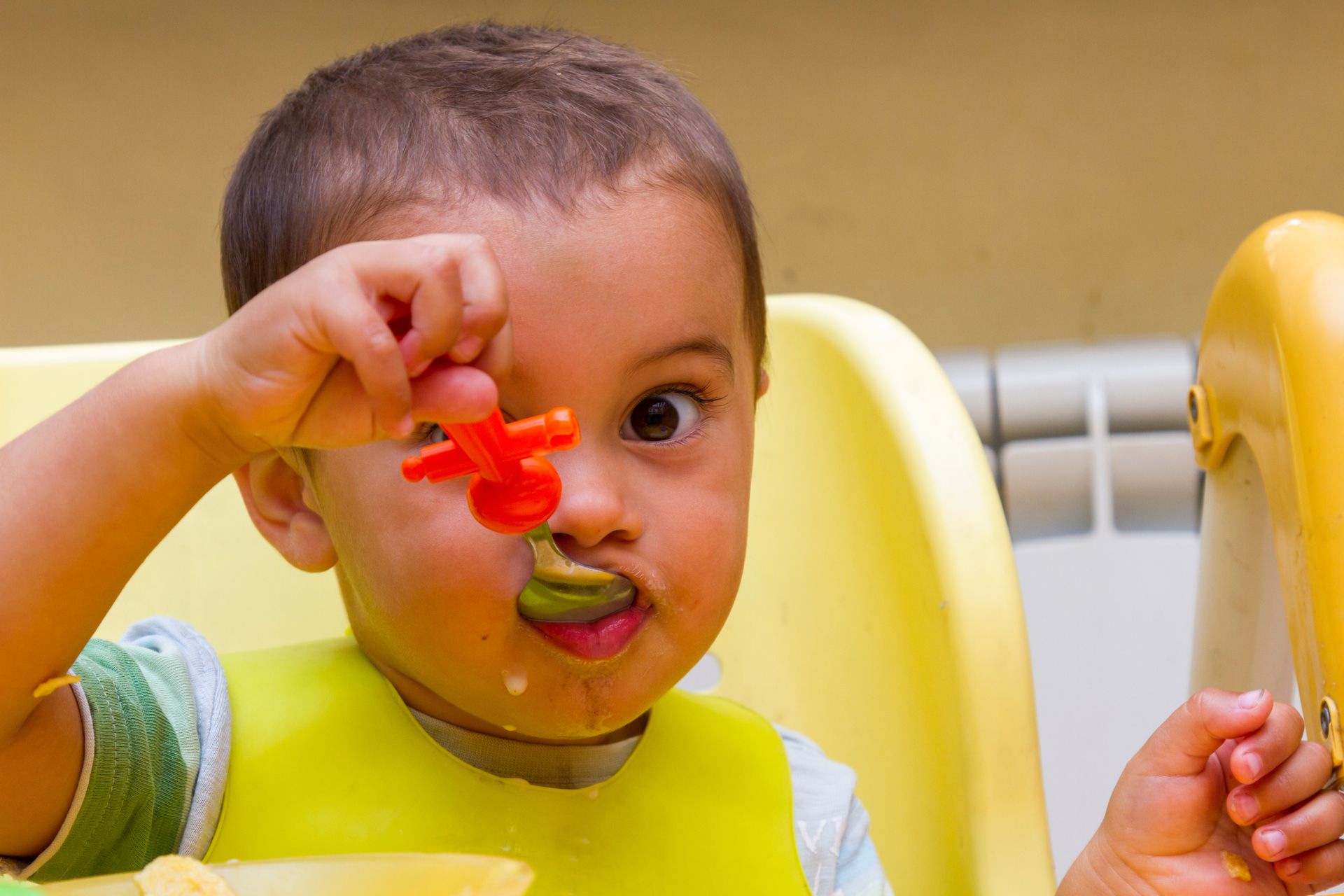 Child in a yellow high chair holding a red toy near their eye and green spoon in their mouth