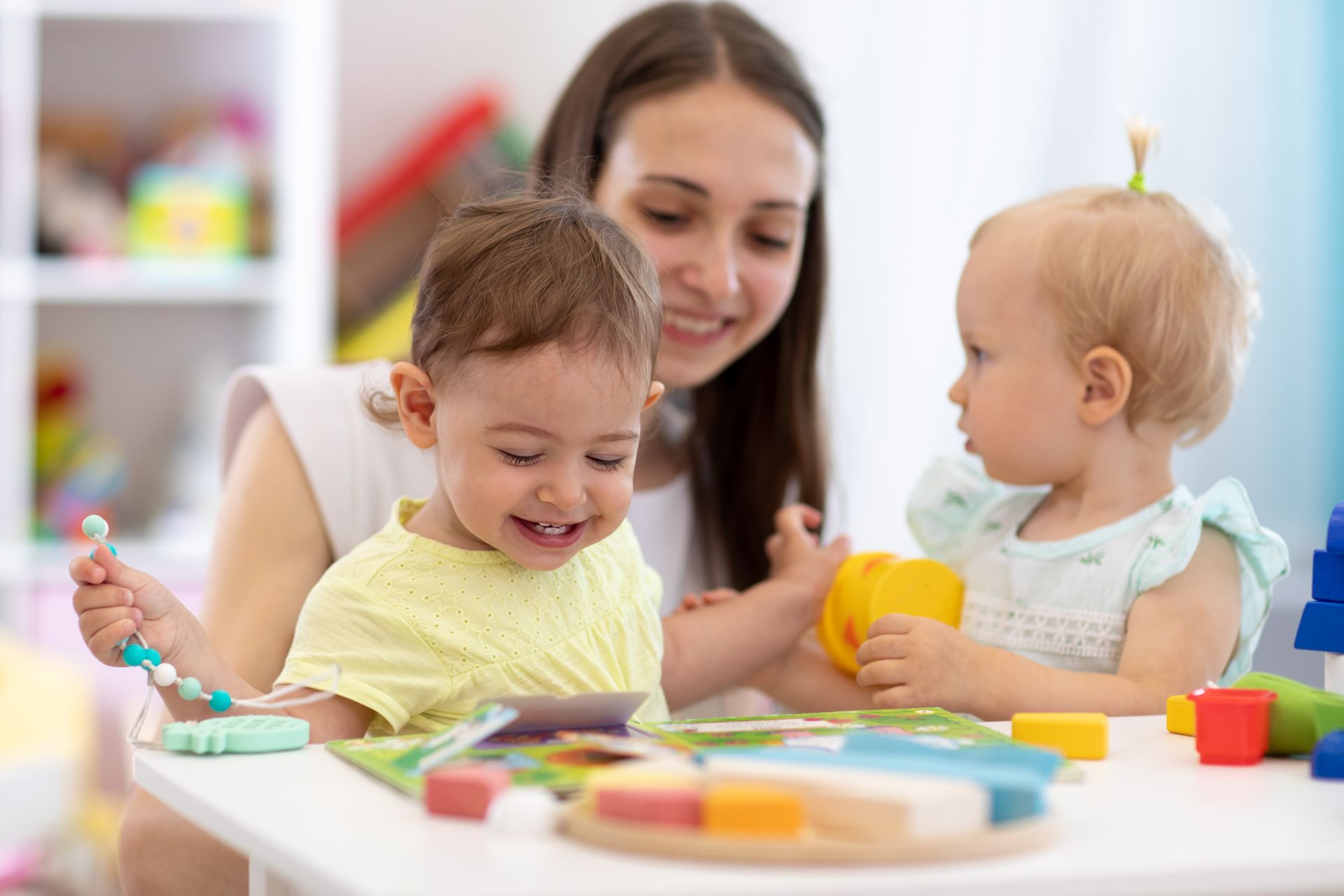 Woman and two toddlers playing with colorful toys at a table in a bright room