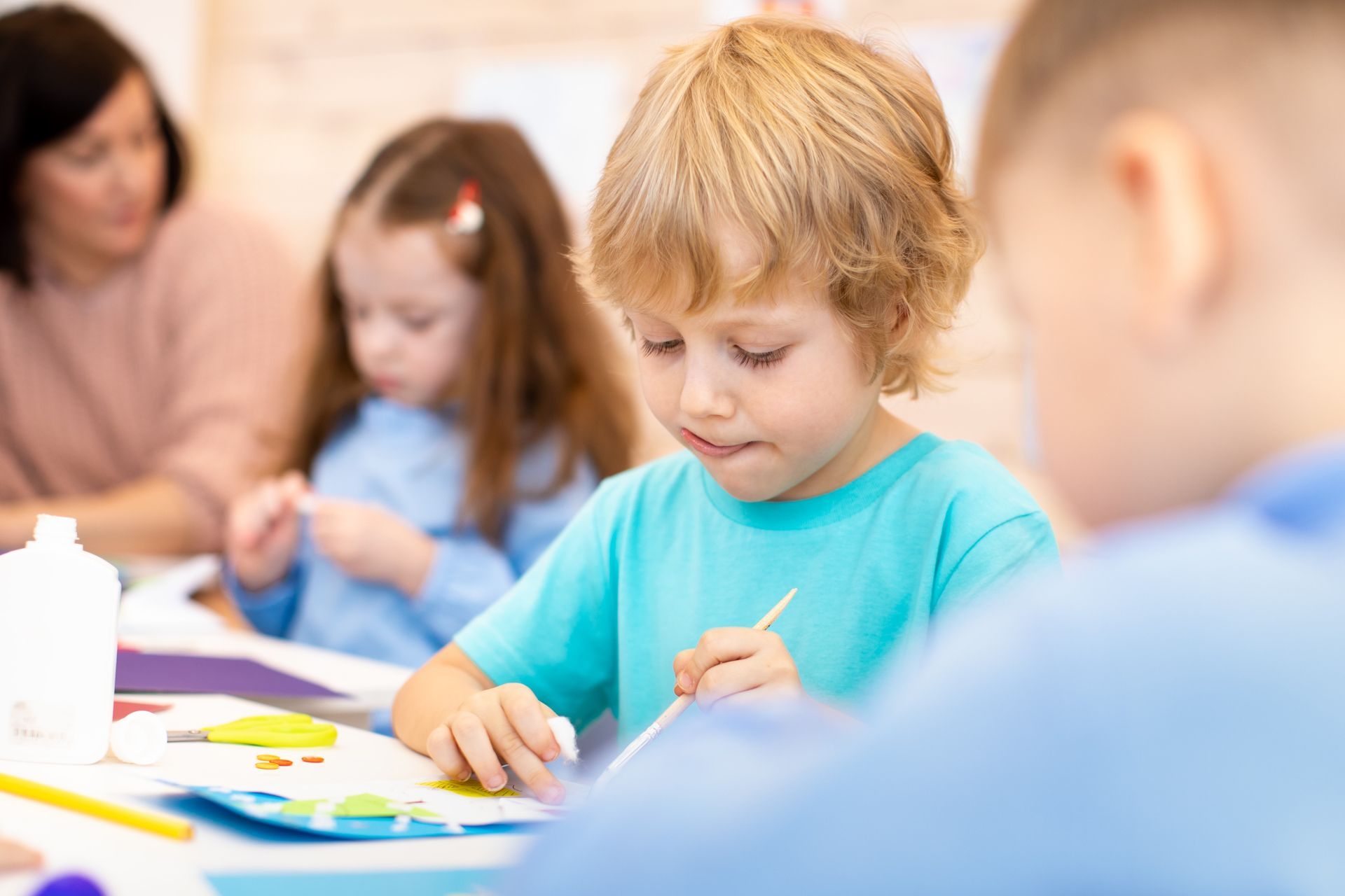 Children painting at a table in a bright classroom, with a teacher helping in the background