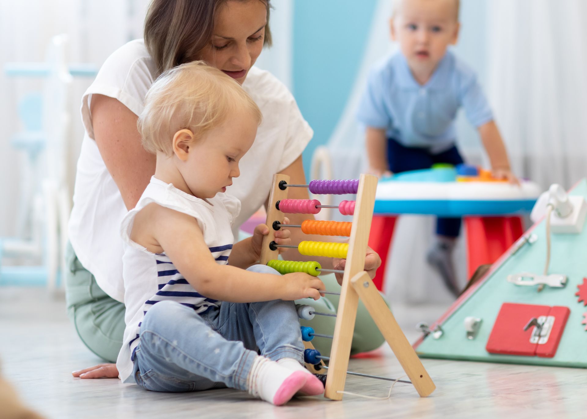 Adult helping toddler with colorful stacking toys on a playroom floor, with another child in the background