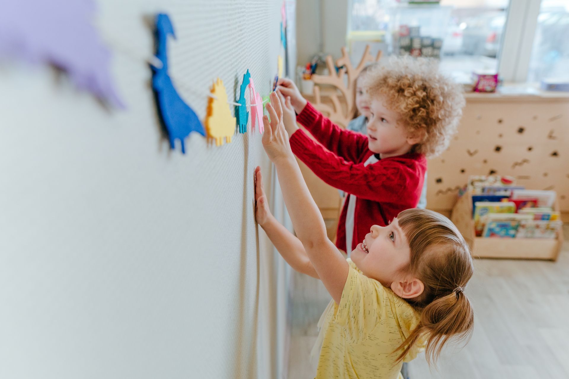 Two children reaching up to place colorful paper birds on a wall display in a bright room