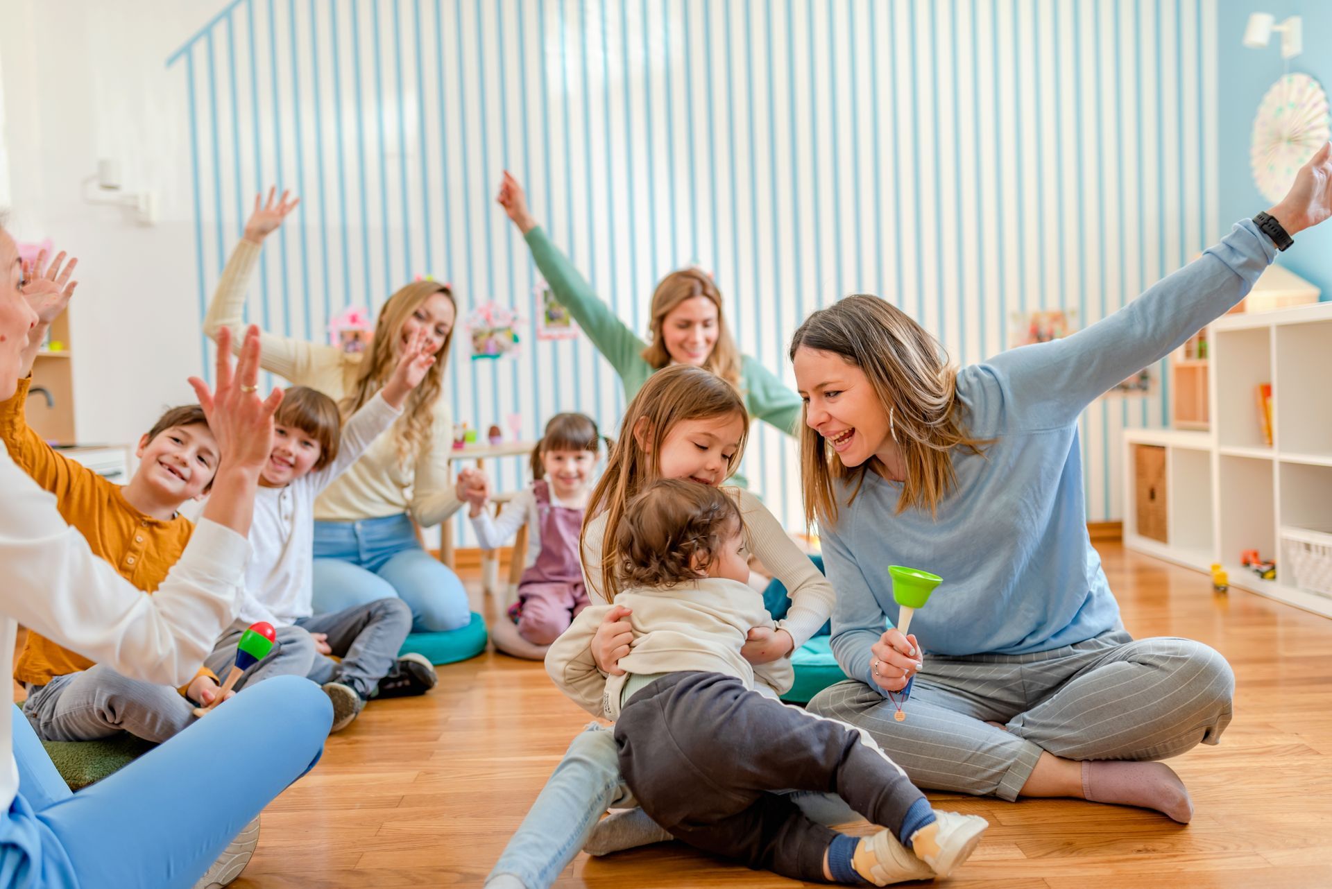 Children and a teacher sitting on the floor, smiling and raising their hands in a bright classroom