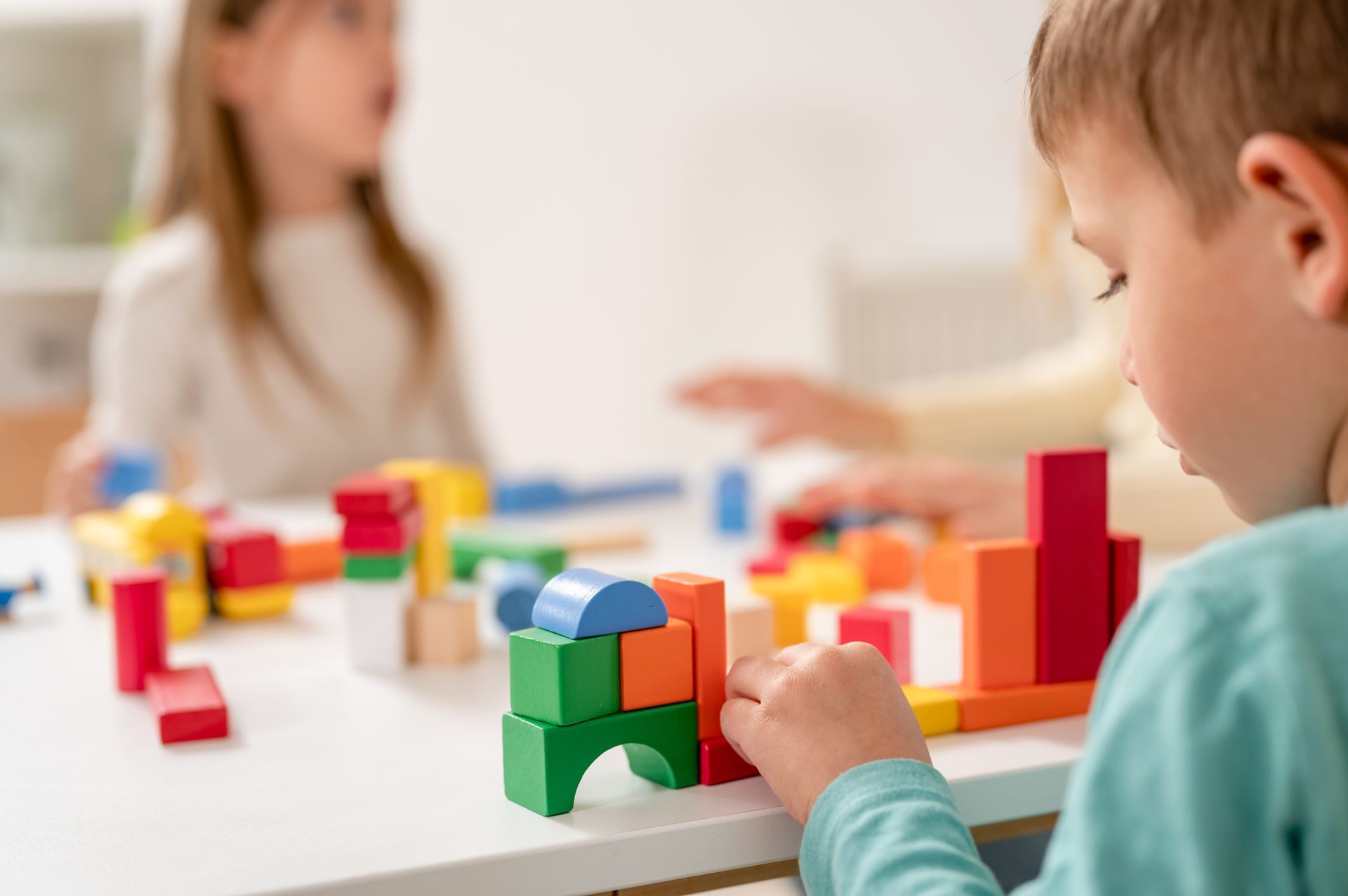 Child playing with colorful blocks at a table while another child is blurred in the background