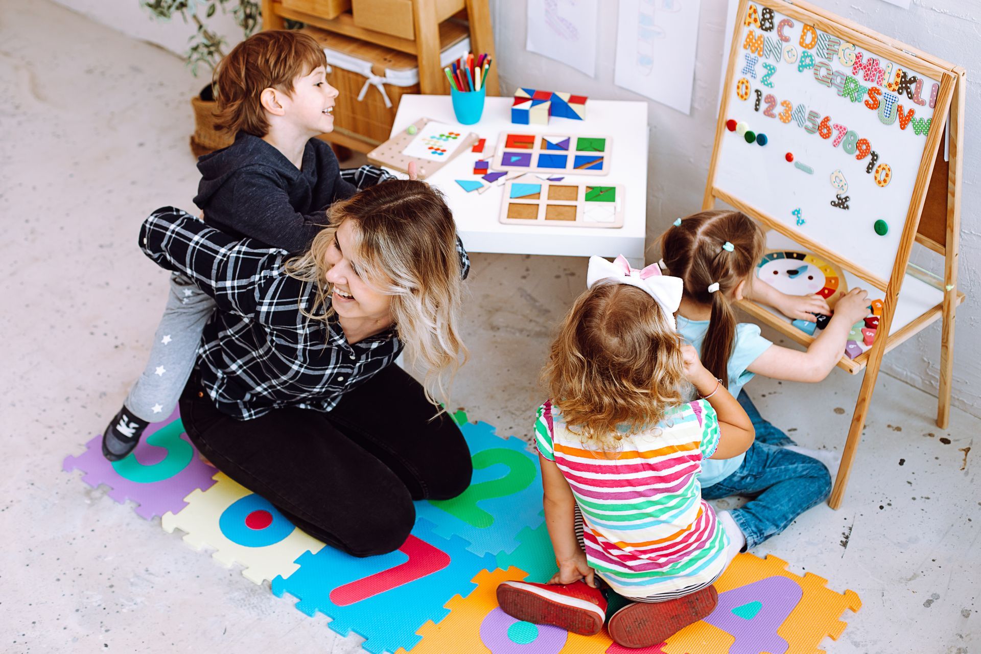 Children painting at a small classroom table, with a teacher on a colorful foam mat nearby