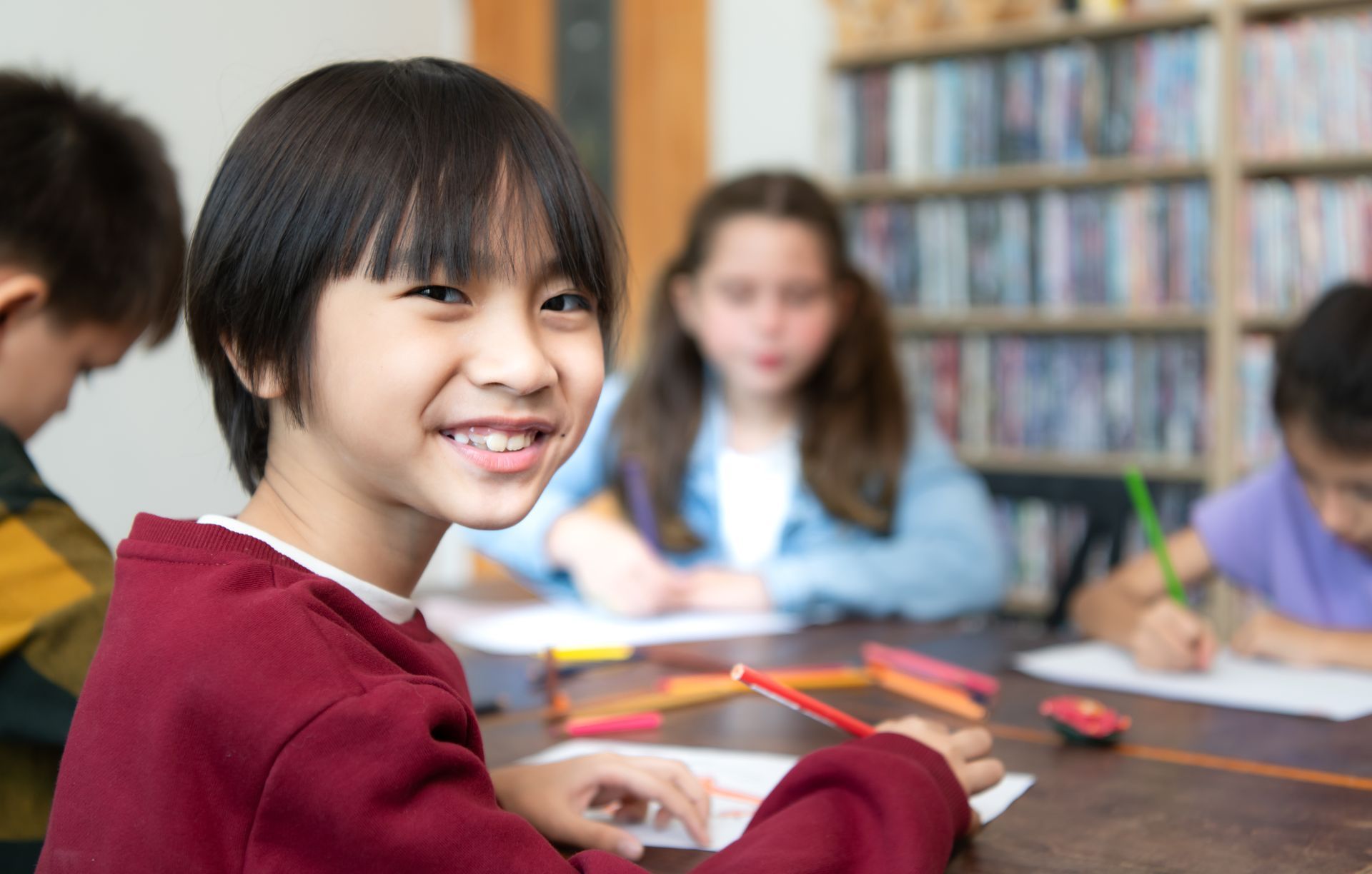 Smiling student in red sweater at classroom table with classmates, pencils, and books in the background