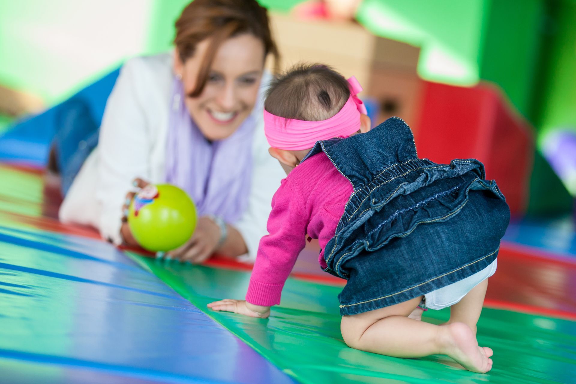Baby crawling on a colorful mat toward a smiling adult holding a green toy ball