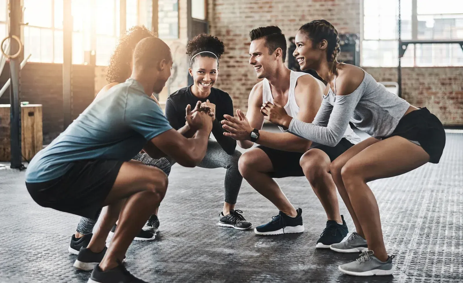 Vier Personen, die in einem Fitnessstudio synchron Kniebeugen machen, lächeln und einander zugewandt.