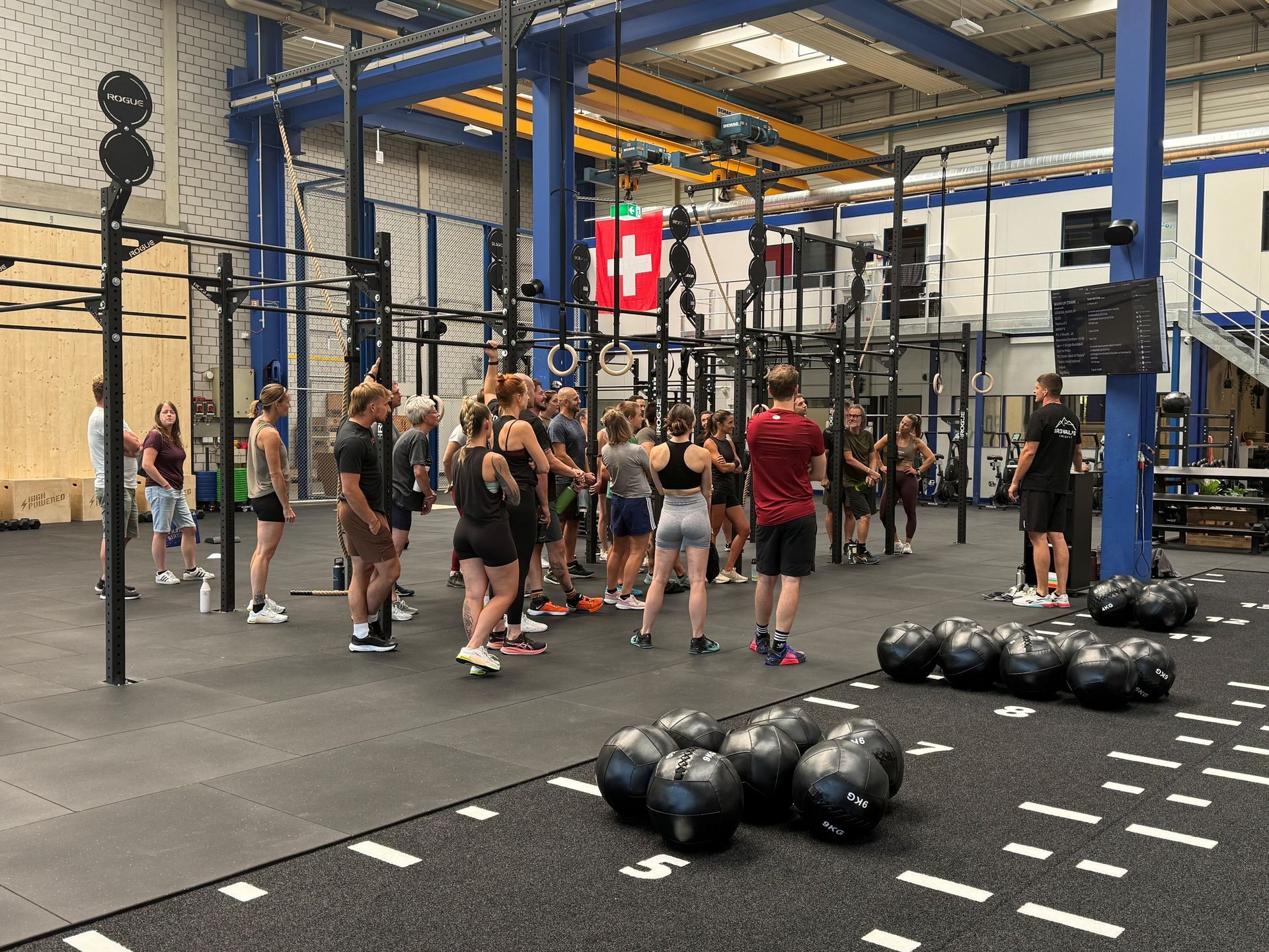 Drei Personen heben Langhanteln in einem Fitnessstudio mit schwarzem Boden und Wellblechwänden.