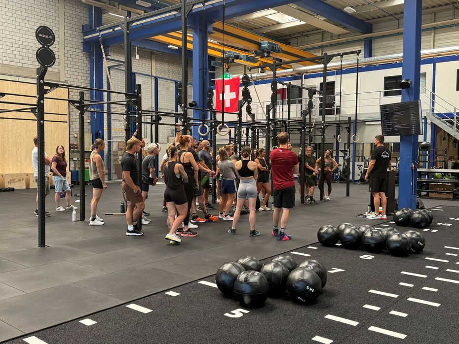 Drei Personen heben Langhanteln in einem Fitnessstudio mit schwarzem Boden und Wellblechwänden.