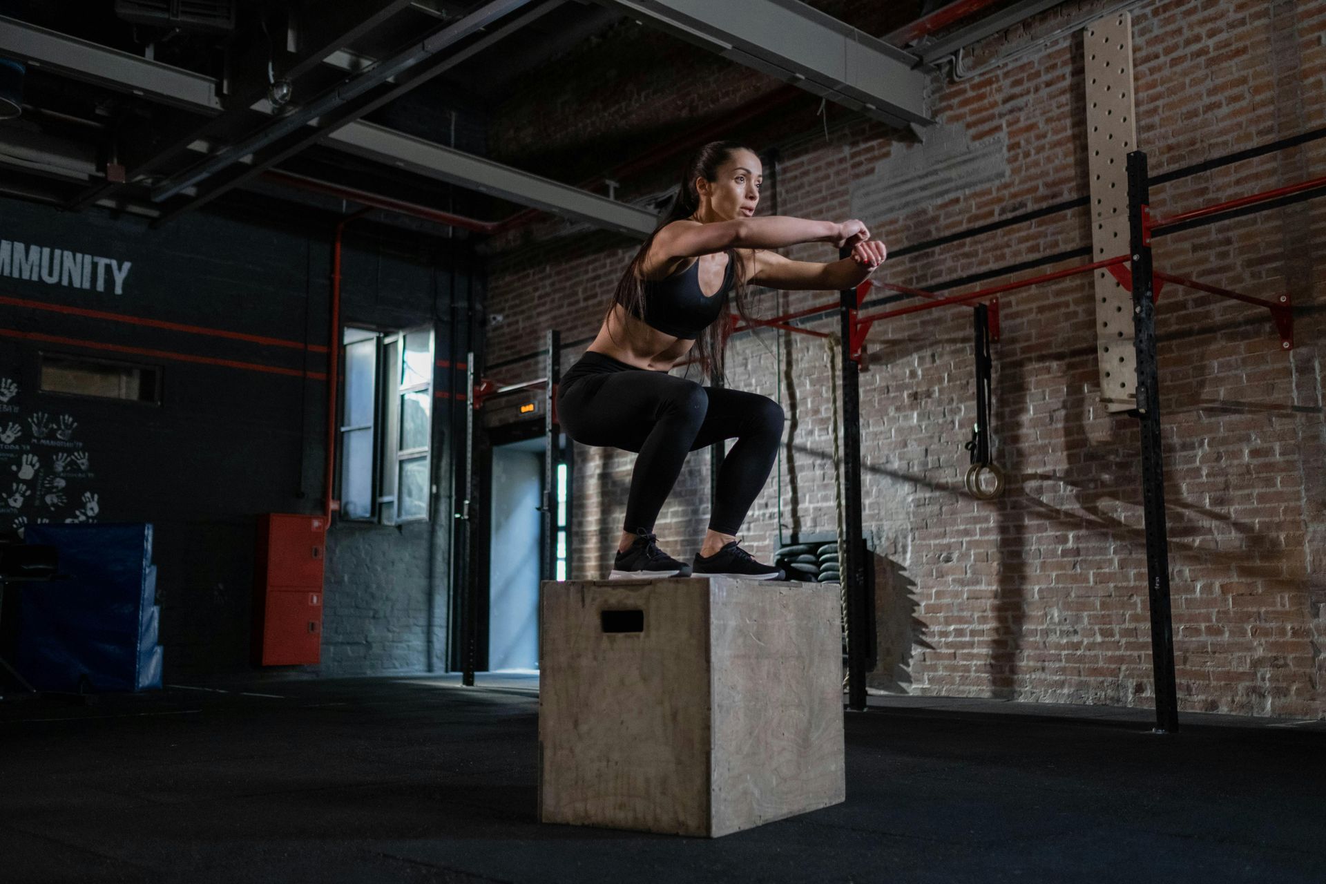 Eine Person führt in einem Fitnessstudio Boxsprünge durch, hält dabei Schlingentrainer fest; die Wände sind aus Ziegelstein, der Boden ist dunkel.