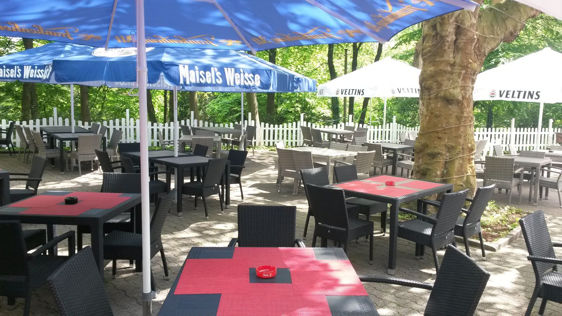 Café im Freien mit Tischen, Stühlen, Sonnenschirmen und weißem Lattenzaun im Schatten.