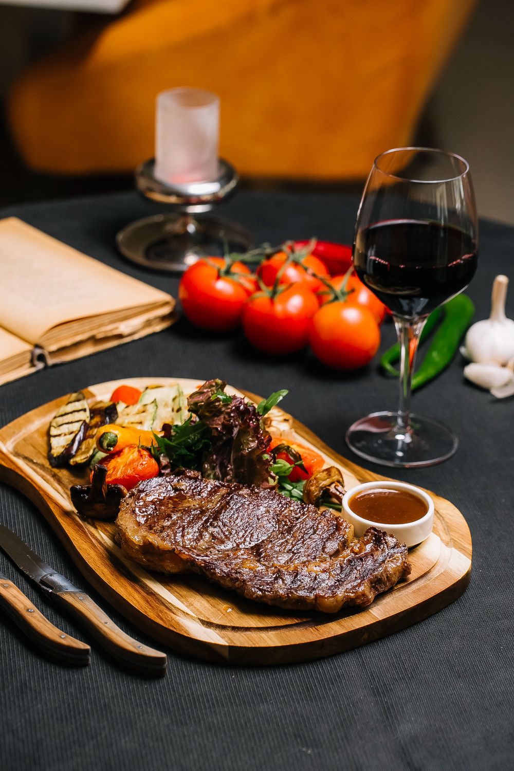 Steak-Dinner auf einem Holzbrett mit geröstetem Gemüse, Tomaten, Wein und einer Kerze.
