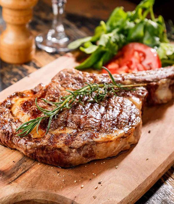Gegrilltes Ribeye-Steak auf einem Holzbrett, garniert mit Rosmarin, Tomaten und Kräutern.